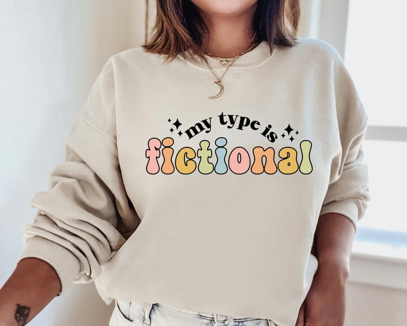 May include: A light beige sweatshirt with the text "my type is fictional" in rainbow-colored letters. The text is in a retro font style.