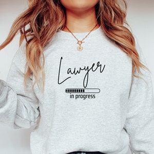 Op de afbeelding: Grijs sweatshirt met de tekst "Advocaat in uitvoering" en een voortgangsbalkgrafiek.