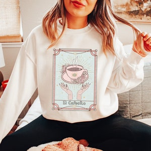 May include: White sweatshirt with a graphic of a coffee cup in a vintage tarot card style. The card is titled "El Cafeito".