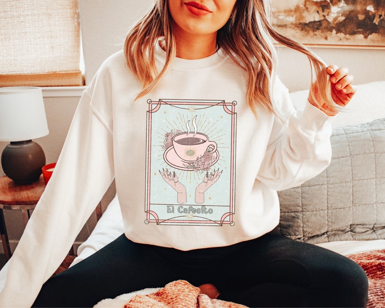 May include: White sweatshirt with a graphic of a coffee cup in a vintage tarot card style. The card is titled "El Cafeito".