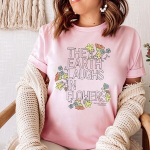 Pode incluir: Uma camiseta rosa com um design floral e o texto "The earth laughs in flowers - Ralph Waldo Emerson".