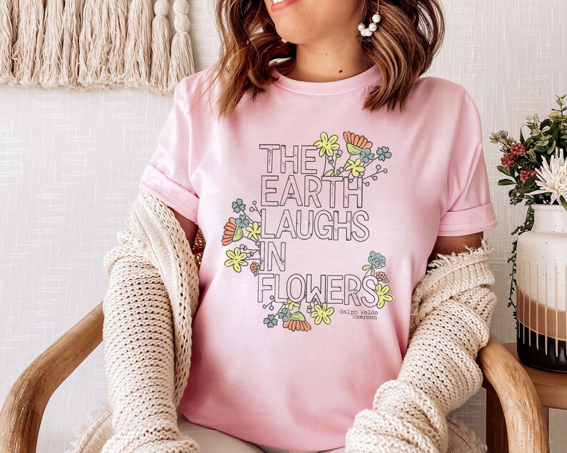 Pode incluir: Uma camiseta rosa com um design floral e o texto "The earth laughs in flowers - Ralph Waldo Emerson".