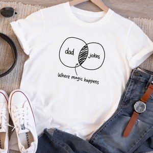 May include: White t-shirt with a black graphic of a Venn diagram with the words "dad" and "jokes" overlapping. The text "where magic happens" is below the diagram.