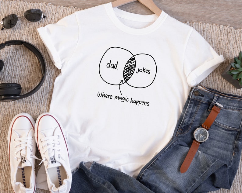 May include: White t-shirt with a black graphic of a Venn diagram with the words "dad" and "jokes" overlapping. The text "where magic happens" is below the diagram.