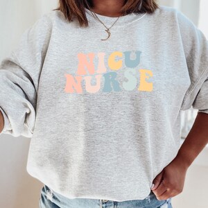 May include: A grey sweatshirt with the words "NICU NURSE" in rainbow colours.