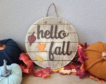May include: A round wooden sign with horizontal lines and the text hello fall in dark brown script. The sign features three small leaf cutouts in burgundy, orange, and yellow. It hangs from a jute twine loop and is surrounded by fabric pumpkins.