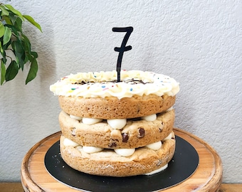 May include: A three-layer chocolate chip cookie cake with white frosting between each layer. The top is decorated with white piped frosting and colorful sprinkles. A black acrylic cake topper in the shape of the number 7 is inserted into the center.