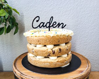 May include: A three-layer cookie cake with white frosting and colorful sprinkles. The cake has a black topper that reads "Caden". The layers are separated by white cream and chocolate chips.