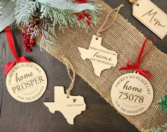 May include: Wooden Christmas ornaments in various shapes, including Texas state outlines and circles. Each ornament is engraved with the phrase "There's no place like home" and a location. A wooden tag with the word "Mom" is also present.