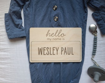 May include: A wooden name plaque with the text "hello my name is" and the name "WESLEY PAUL" engraved in a bold font. The plaque is placed on a blue onesie with button details, next to a pacifier clip.