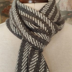 May include: A gray and white woven scarf with a geometric pattern is draped over a mannequin. The scarf has a fringed edge and is made of a soft, textured material. The scarf is a neutral color and would be suitable for any outfit.