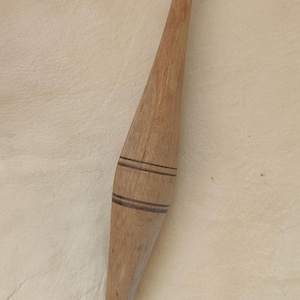 Può includere: Un fuso di legno dal design affusolato. Il fuso è marrone chiaro con due bande scure vicino al centro. Le estremità sono appuntite. Probabilmente viene utilizzato per filare o altre arti tessili.