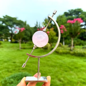 Puede incluir: Una piedra preciosa esférica translúcida de color rosa pálido montada sobre un soporte de metal plateado. El soporte cuenta con una base vertical, una varilla central que atraviesa la esfera y un brazo curvo que sostiene la varilla.