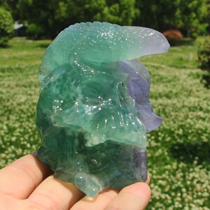 May include: A carved fluorite sculpture in shades of green and purple, depicting a stylized skull with a textured surface. The sculpture is held in a hand, with a blurred green background.