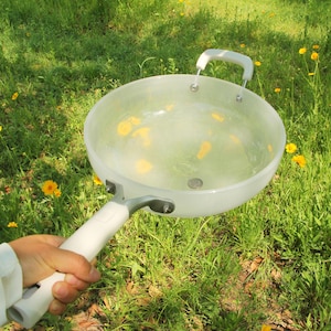 May include: A clear, round-bottomed pan with a white handle and a smaller, curved handle. The pan is held over a grassy area with yellow flowers. The pan appears to be filled with water and yellow flower petals.