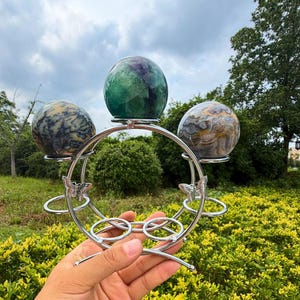 May include: A silver-toned metal display stand featuring a circular frame with butterfly accents, holding three polished stone spheres. The top sphere is green and purple, while the two side spheres feature marbled patterns in blue, grey, and tan.
