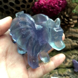May include: A translucent, blue and purple carved crystal figurine of a mythical creature with wings, resembling a bear or a similar animal. The figurine is held in a hand, with details of the wings and body clearly visible. The crystal has a polished finish.
