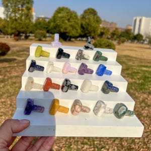May include: A collection of decorative bottle stoppers in various colors and materials, including green, black, clear, pink, purple, and blue. The stoppers are displayed on a white stepped platform, showcasing their unique designs and textures.
