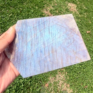 May include: A rectangular, polished labradorite stone with a blue and white iridescent sheen. The stone has a natural, textured surface with visible veining. The edges are cut at an angle, and the stone is held against a backdrop of green grass.
