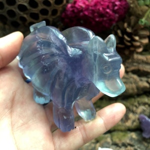 May include: A translucent, blue and purple carved crystal figurine of a mythical creature with wings. The figurine has a bear-like body with a long snout and small ears. The wings are spread out, and the crystal has a smooth, polished finish.