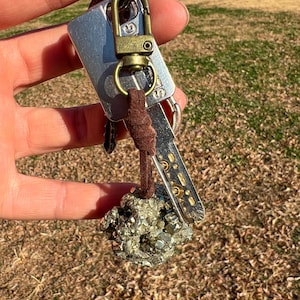 以下が含まれることがあります： 黄鉄鉱の石のペンダントが付いたキーリング。キーリングは、シルバーのタグ、真鍮色の留め金、茶色のスエードコードが特徴です。黄鉄鉱は金属的で金色の外観をしています。鍵はキーリングに取り付けられています。