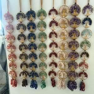 May include: A collection of wire-wrapped pendants featuring a tree of life design. Each pendant is a different color, with gemstones in shades of pink, brown, blue, green, red, white, and purple. The pendants hang from gold-colored chains.