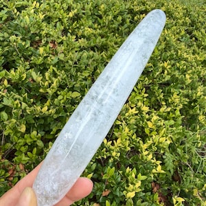Pode incluir: Uma varinha de cristal de quartzo transparente, longa e cilíndrica, com uma superfície lisa e polida. A pedra apresenta inclusões internas naturais, fraturas e manchas brancas nubladas por todo o seu corpo translúcido, terminando em ponta.