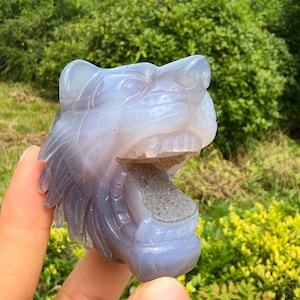 May include: A translucent, light blue-grey carved stone sculpture in the shape of a roaring tiger head. The carving features detailed facial contours, including ears, eyes, and teeth, with a rough, textured, grey mineral deposit inside the open mouth.