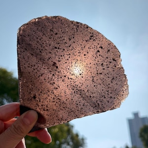 Puede incluir: Una piedra translúcida, de forma irregular, de color marrón rojizo con una textura porosa. El sol brilla a través de la piedra, creando un punto brillante en el centro. La piedra se sostiene contra un cielo azul.