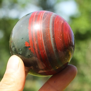 Peut inclure: Une sphère de pierre précieuse polie avec un mélange de couleurs rouge foncé et vert foncé. La sphère présente un motif tourbillonnant de rayures rouges et blanches. La pierre est tenue par les doigts d'une personne.