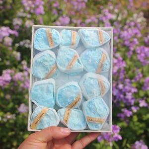 May include: A white box filled with twelve light blue aragonite stones. Each stone is wrapped in white tissue paper and secured with a small label that reads "ARAGONITE". The background is a blur of purple flowers.