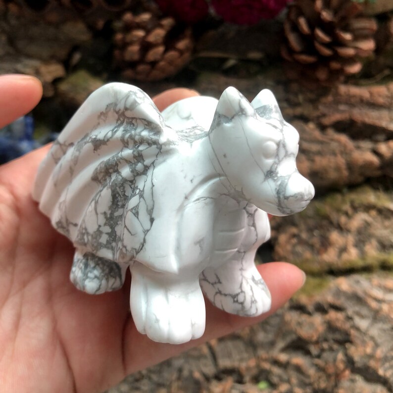May include: A white marble dragon figurine with gray veining. The dragon has detailed wings, a textured body, and is carved with intricate features. The figurine is held in a hand, showcasing its size and craftsmanship.