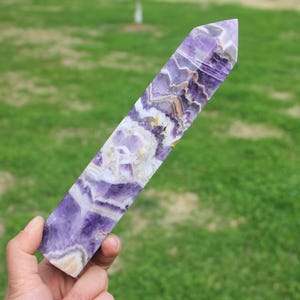Puede incluir: Una punta de cristal de amatista pulida, con una mezcla de tonos morados, blancos y beige. La forma alargada y rectangular se estrecha hasta un extremo puntiagudo. El cristal se sostiene sobre un fondo de hierba verde.