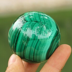 May include: A polished, spherical malachite stone. The gemstone displays a vibrant green color with swirling patterns of lighter and darker green hues. The surface is smooth and reflective, with a glossy finish.