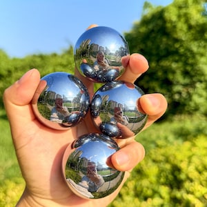 Puede incluir: Cuatro bolas metálicas esféricas pulidas con una superficie altamente reflectante similar a un espejo. Cada esfera captura un reflejo distorsionado y gran angular del entorno exterior, incluyendo árboles, edificios y una persona que las sostiene.