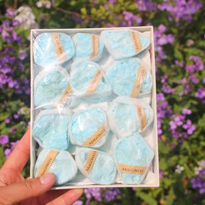 May include: A box of light blue aragonite stones, each wrapped in clear plastic and labeled with a brown sticker. The stones have a rough, textured appearance. The box is held in front of a background of purple flowers.