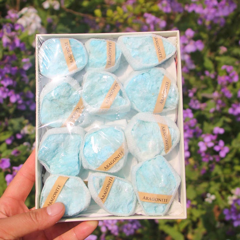 May include: A box of light blue aragonite stones, each wrapped in clear plastic and labeled with a brown sticker. The stones have a rough, textured appearance. The box is held in front of a background of purple flowers.