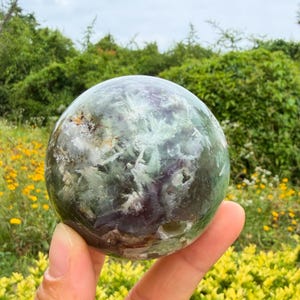 May include: A polished spherical stone featuring a translucent green and purple color palette with internal white fibrous inclusions and mineral deposits. The surface is smooth and reflective, showcasing natural variations in the mineral structure.