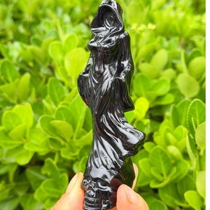 May include: A black figurine of a hooded figure, possibly the Grim Reaper, with a skull base. The figure is carved with flowing robes and a detailed hood. The statue is held against a backdrop of green foliage.