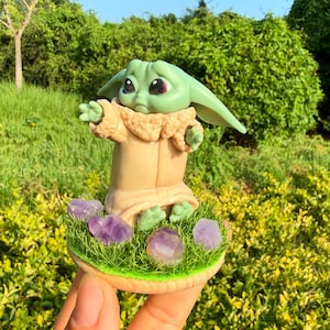 May include: A small green creature figurine with large dark eyes and pointed ears, wearing a tan robe with a textured collar. The figure stands on a circular base covered in artificial green grass and decorated with several small purple amethyst crystals.