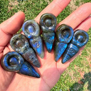 Peut inclure: Six figurines en labradorite sculptées, chacune avec une tête arrondie et un corps allongé. Les pierres présentent des reflets bleus et verts irisés. Les figurines sont tenues dans une main sur un fond d'herbe verte.