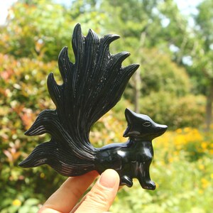 May include: A black fox figurine with a large, stylized tail. The fox is standing, with its tail fanned out behind it. The figurine is made of a glossy, dark material, and is held in a person's hand.