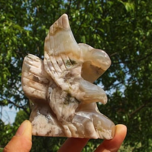 May include: A carved stone eagle sculpture. The eagle is depicted with outstretched wings and detailed feathering. The sculpture features a mix of beige, white, and light brown colors with some green inclusions. The sculpture is held in a hand.
