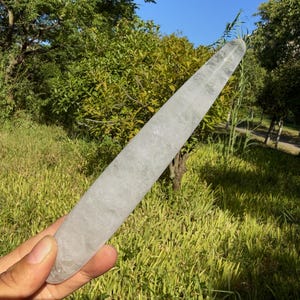 Könnte beinhalten: Ein langer, schlanker Stab aus klarem Quarzkristall mit einer glatten, polierten Oberfläche und einer spitz zulaufenden Form. Der Kristall ist durchscheinend mit sichtbaren inneren Einschlüssen und natürlichen Rissen in blasser, eisweißer Farbe.