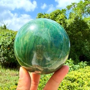 May include: A polished, spherical stone with a smooth, glossy surface featuring swirling patterns of deep forest green, emerald, and lighter lime green hues, reflecting light across its rounded, solid form held in a hand.