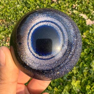 Puede incluir: Un objeto esférico con un acabado liso y brillante que presenta un centro azul profundo rodeado de anillos concéntricos de purpurina plateada y azul oscuro. La superficie es reflectante y muestra un reflejo circular oscuro en el centro.