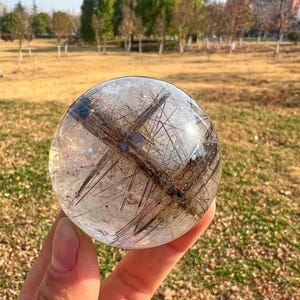 May include: A clear crystal sphere with dark brown and black inclusions. The orb is held in a hand against a blurred background of grass and trees. The crystal has a polished, reflective surface.