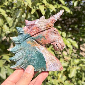 May include: A carved stone unicorn head sculpture. The sculpture features a detailed mane and horn, with colors of green, red, and white. The sculpture is held in a hand against a background of green foliage.