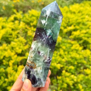 May include: A polished fluorite crystal tower featuring a hexagonal prism shape with a pointed top. The stone displays a blend of translucent green and deep purple hues, characterized by internal white feathery inclusions and natural mineral fractures.