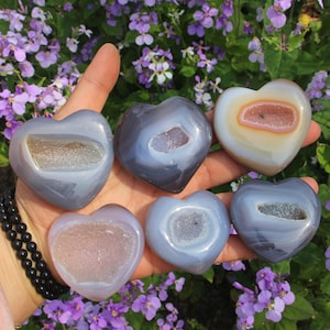 May include: Six heart-shaped agate stones in shades of gray, blue, and cream are displayed. Each stone has a geode center. The stones are held in a hand against a backdrop of purple flowers.